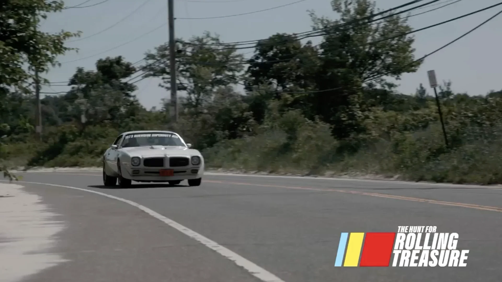 White Pontiac Trans Am driving toward the camera on a sunlit road